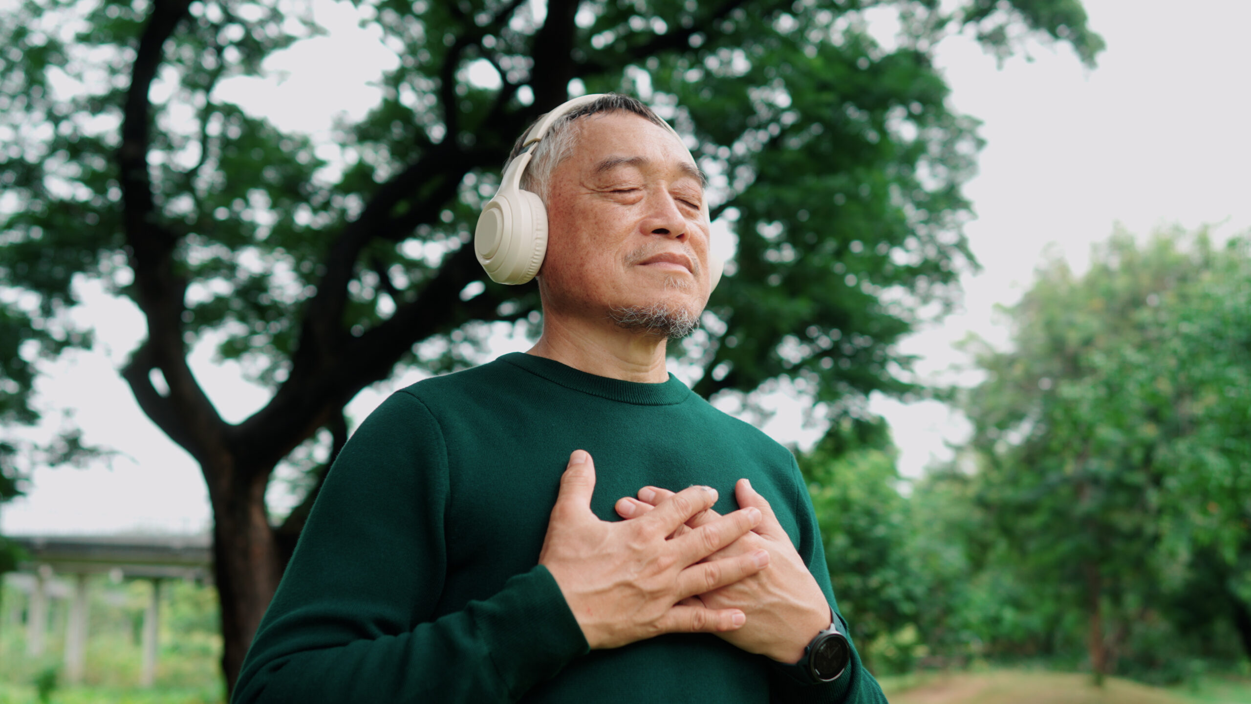 Elderly Asian man meditating under a tree with wireless headphones, calm and refreshed.
