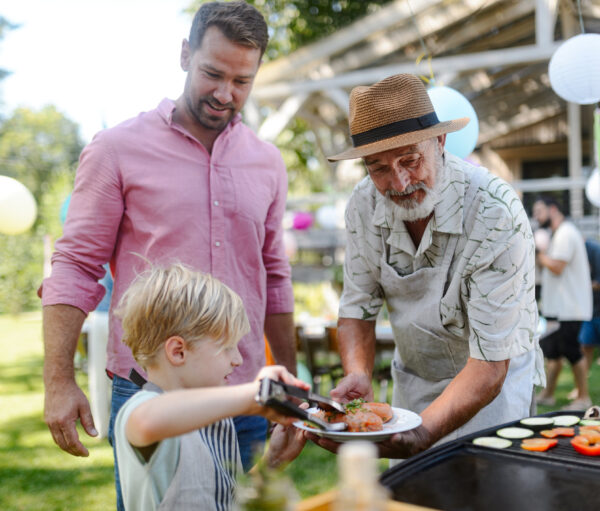 Father, grandfather and son grilling together at a garden bbq party. Three generations of men at summer family garden party.